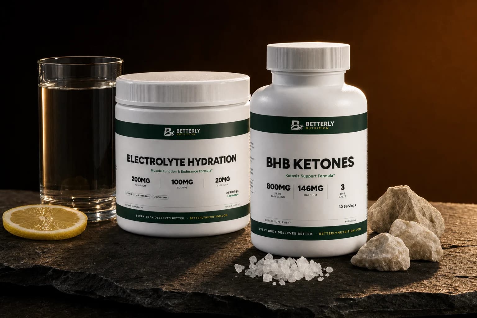 BHB Ketones bottle beside an Electrolyte Hydration Lemonade tub on dark slate with a glass of water, lemon slice, sea salt and pale stones