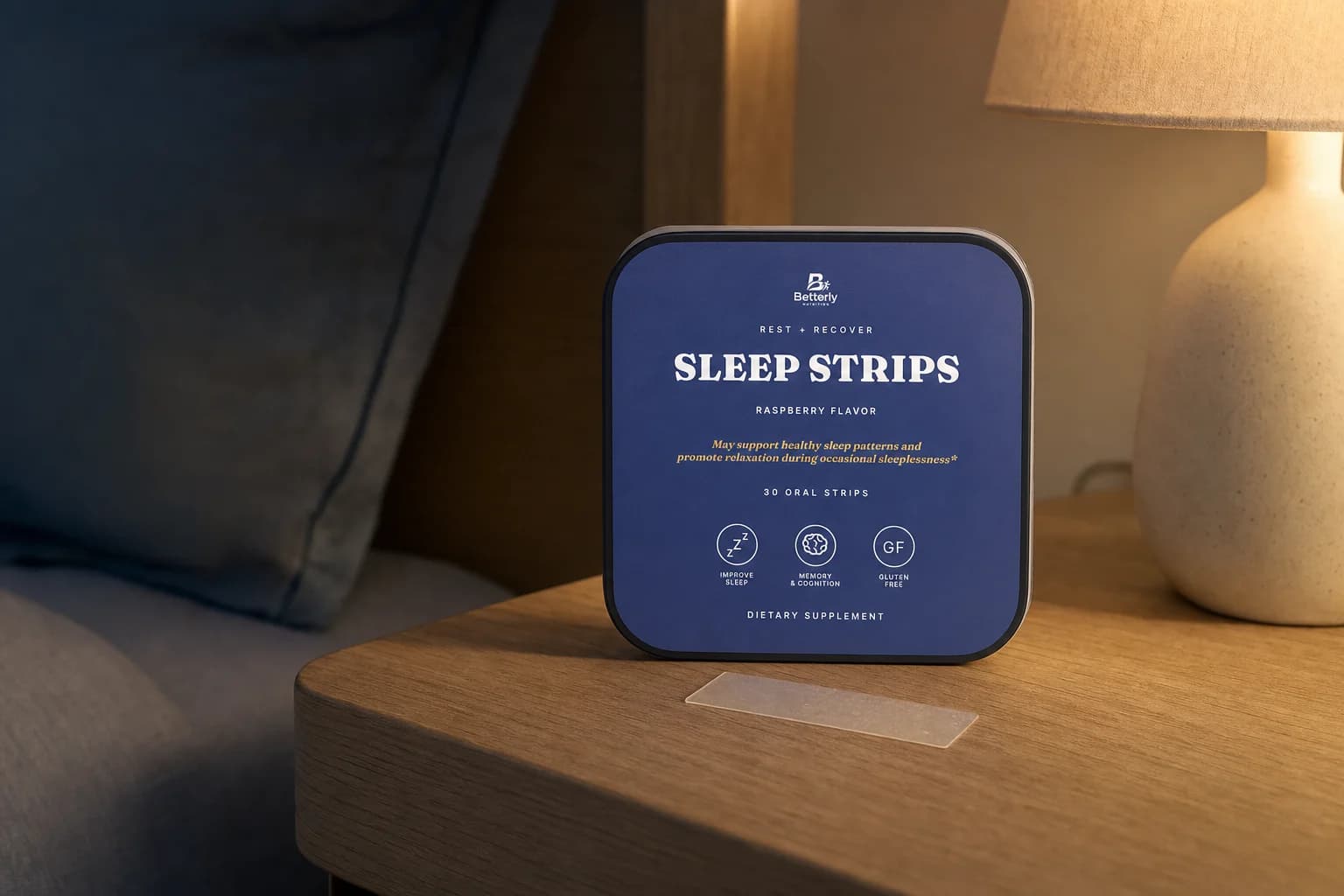 Sleep Strips tin standing on a pale-oak nightstand with one translucent dissolvable strip in front, in warm bedside lamplight